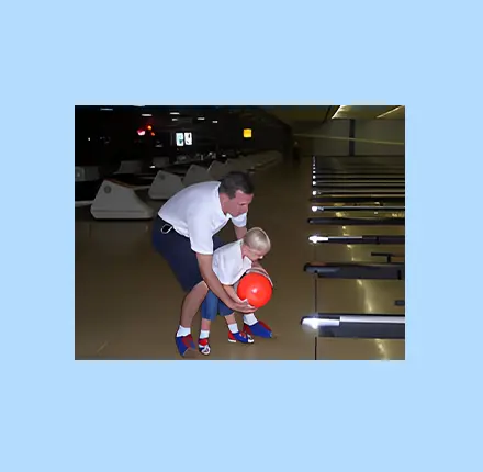 A man helping a child learn to bowl at a bowling alley.