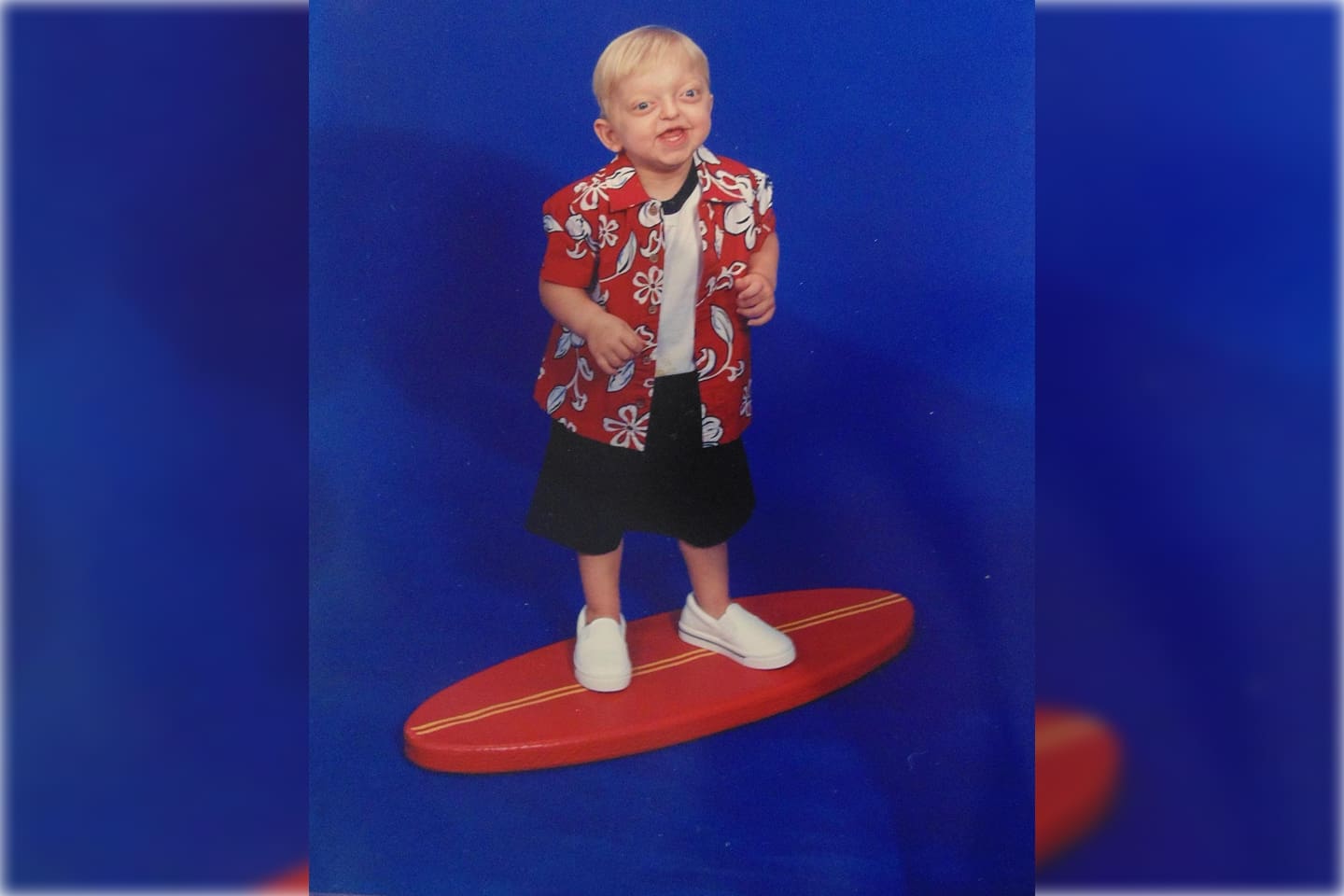 A young child smiling while standing on a red skateboard against a blue background.